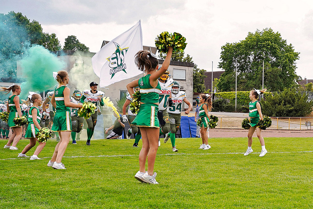 Eine Gruppe von Cheerleadern in grünen Uniformen begrüßt American-Football-Spieler, die aus einer Rauchwolke kommen, mit einer großen Flagge und Pompons in der Hand auf einem Sportfeld.