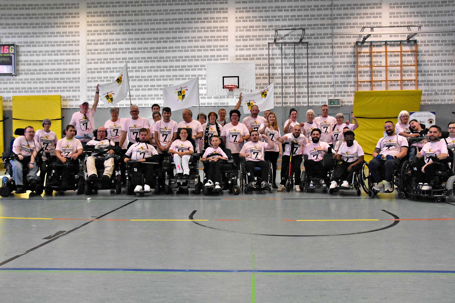 Gruppe von Menschen in Rollstühlen und ihren Unterstützern posieren in einer Sporthalle, alle tragen rosa T-Shirts mit dem Aufdruck „Rollstuhlsport“. Einige halten Fahnen. Im Hintergrund sind Sportgeräte und ein Basketballkorb zu sehen.